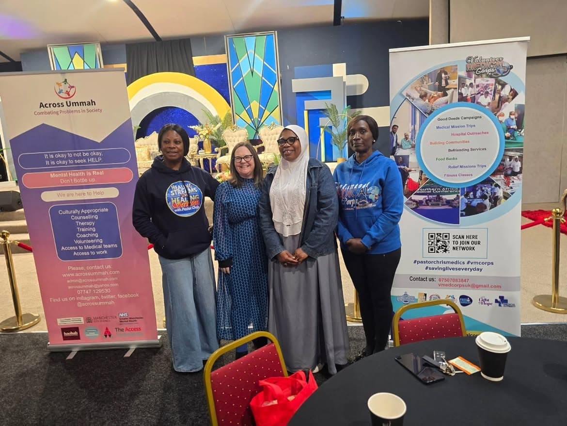 Four women standing together and smiling between two partner organisation pull-up banners at an indoor event.