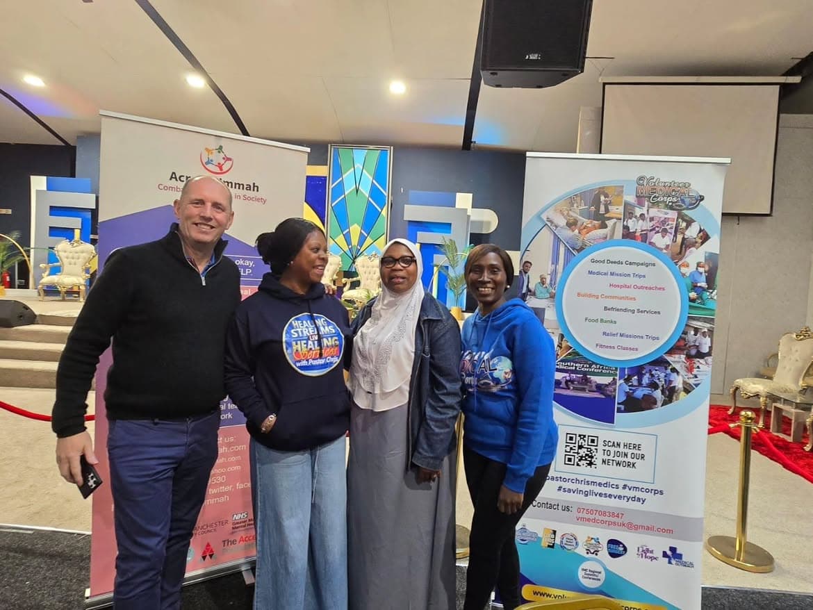 Four people standing together and smiling between two partner organisation banners at an indoor event.