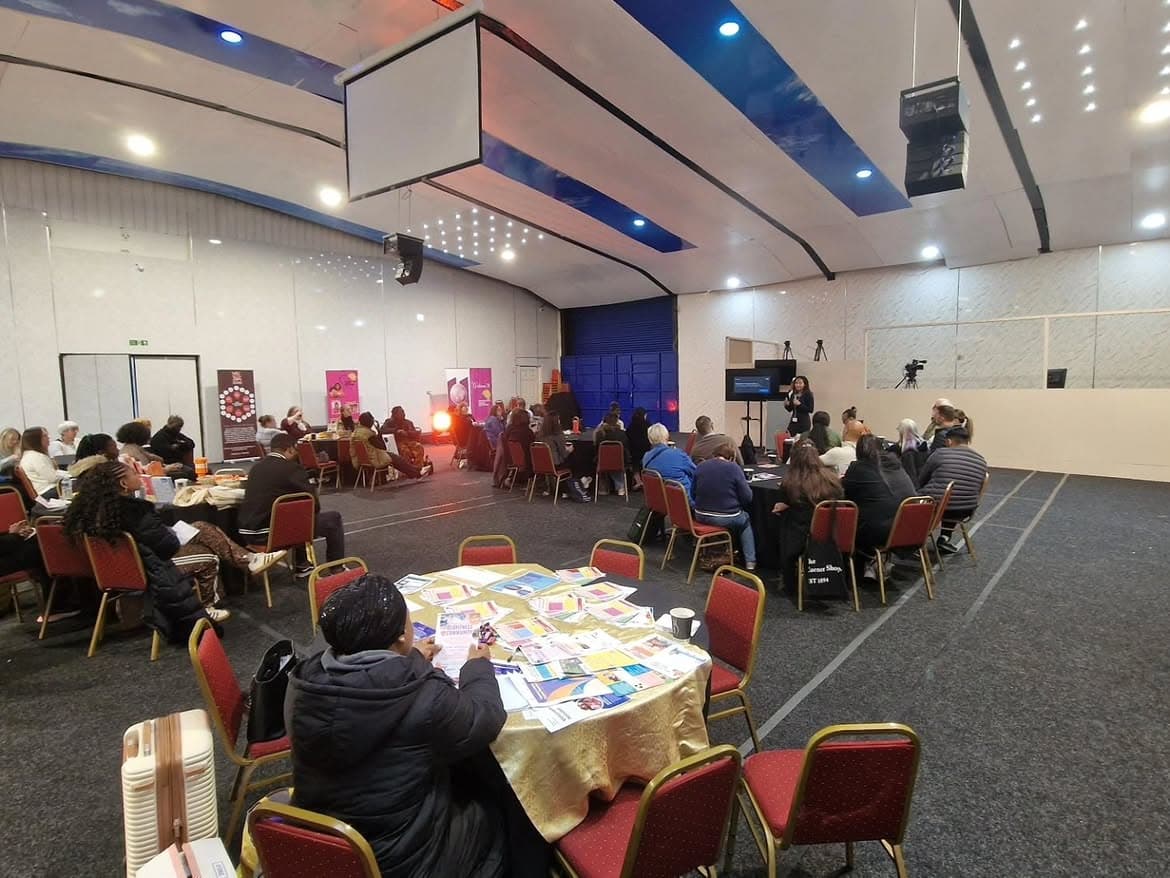 A community hall with people seated at round tables, one table in the foreground spread with printed leaflets and information sheets.